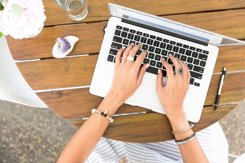 Hands typing on a laptop computer on a brown wood table with various items next to it.