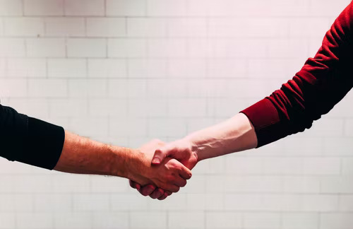 Two hands, one in a black sleeve, the other in a red, shaking hands in front of a white wall.