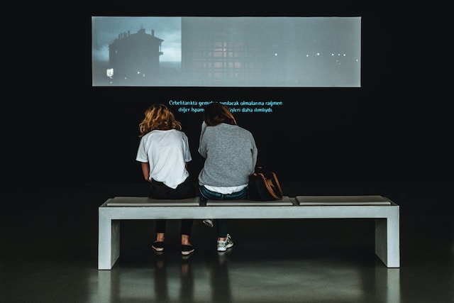 Doi oameni se uită la un videoclip pe un ecran mare cu subtitrări. 