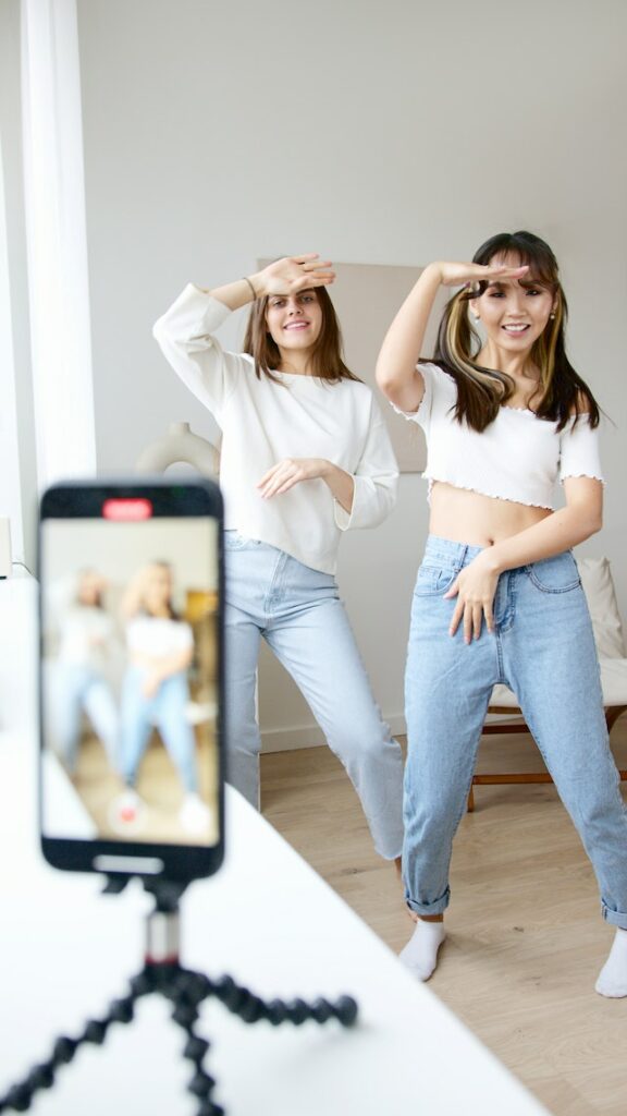 Două femei cu bluze albe și blugi din denim dansează în fața unui telefon pentru un TikTok.