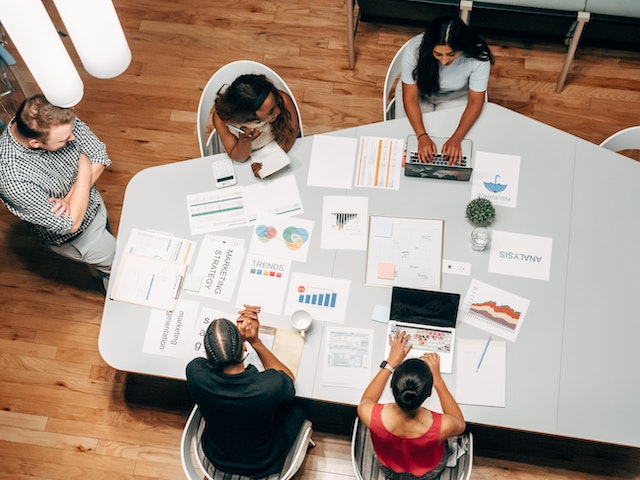 Vista aérea de um grupo de pessoas ao redor de uma mesa com papéis, computadores e materiais de projeto.