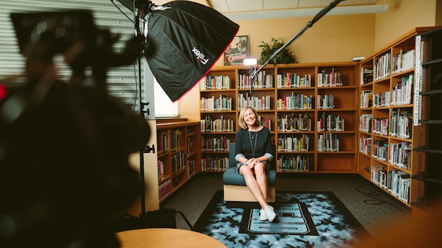 Une femme dans une bibliothèque, assise devant un appareil photo. 