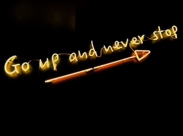 Um letreiro de neon dizendo "Go up and never stop" (Suba e nunca pare) com uma seta iluminada embaixo. 