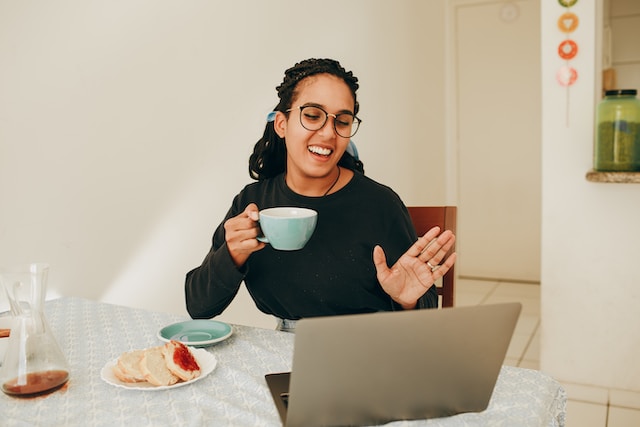 O femeie așezată la o masă în fața laptopului ei, în timp ce ține în mână o ceașcă de cafea. 