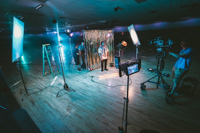 A man standing in front of cameras with string lights behind him. 