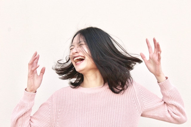 Una donna con un maglione rosa si scompiglia i capelli e ride. 