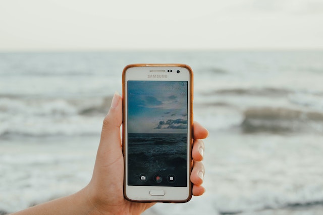 A person holding a white Samsung Android smartphone taking a photo of the ocean.