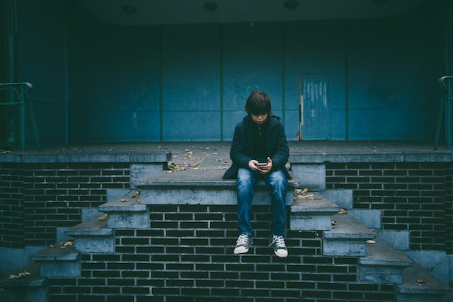 Un băiat așezat pe treptele unei clădiri vechi în timp ce își folosește telefonul. 