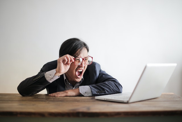 Un homme assis, tenant ses lunettes, et hurlant sur son PC.