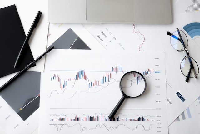 An amplifying glass, documents containing market projections, a pair of eyeglasses, and a pen on a table.