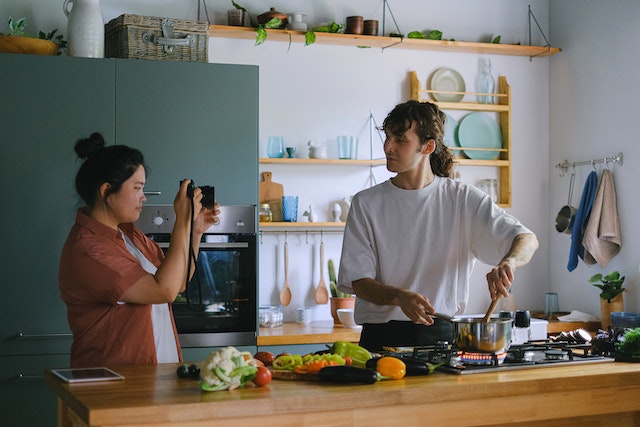 Una mujer graba a un hombre con una cámara digital mientras cocina.