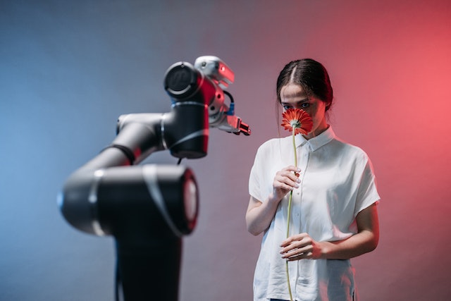 A woman smelling a flower while staring at a robot.