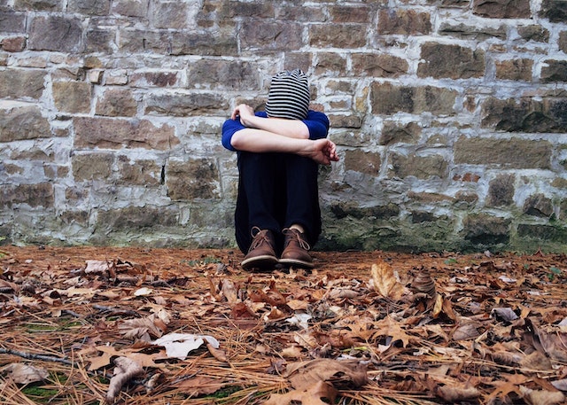 A boy sitting on the floor resting on a brick wall with his head bowed.