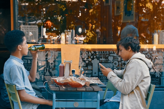 Twee jongens eten buiten een maaltijd, terwijl één jongen op zijn telefoon aan het surfen is. 