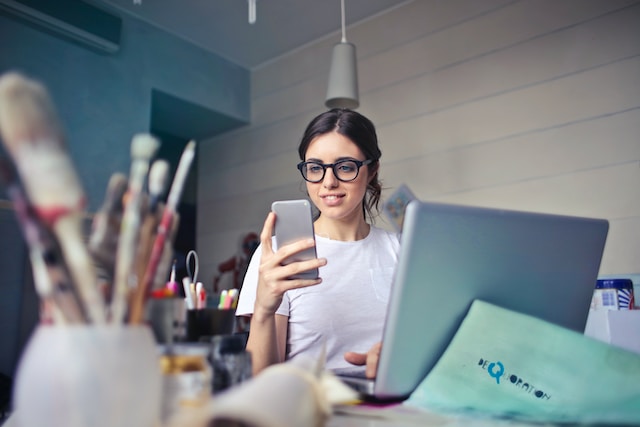 Una mujer con camisa blanca que trabaja en su PC y sonríe al ver algo en su teléfono. 