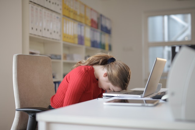 Una donna seduta su una sedia nel suo ufficio e che appoggia la testa al computer portatile sul suo tavolo.