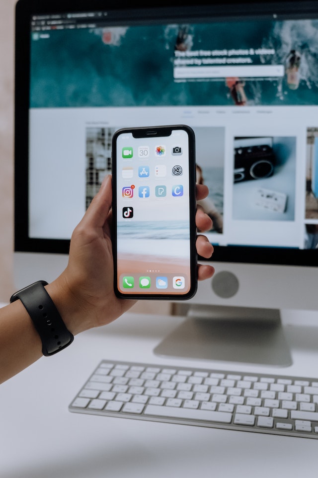 Une personne tenant un smartphone avec l'application TikTok ouverte devant un iMac.