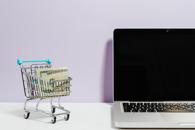 A shopping cart with dollar bills beside a laptop.