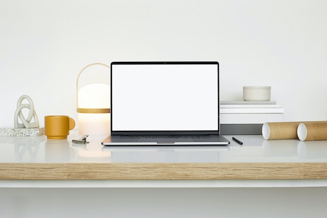 Un MacBook, un bolígrafo y una taza sobre una superficie blanca.