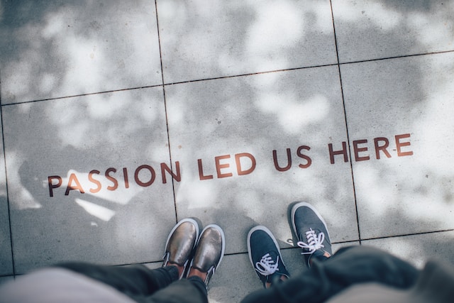 Two people standing beside a sign on the sidewalk that reads, “Passion led us here.”