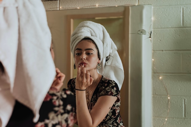 A woman with a towel around her head putting on makeup in front of a mirror.