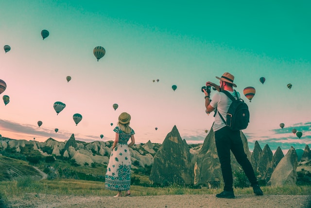A man taking photos of a woman in front of a mountain landscape and hot air balloons. 