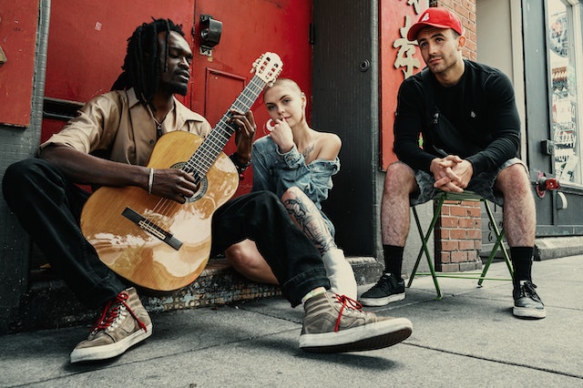 A TikTok musician with a guitar sitting on a stoop with two people sitting next to him and listening.