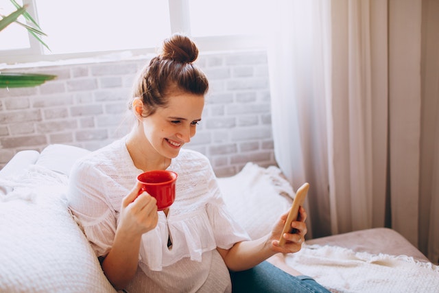 Een dame met een kopje in haar hand, glimlachend en scrollend door haar TikTok-pagina op haar telefoon.