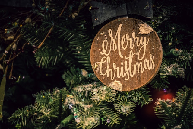 A wooden Christmas ornament with the words “Merry Christmas” hangs on a tree. 