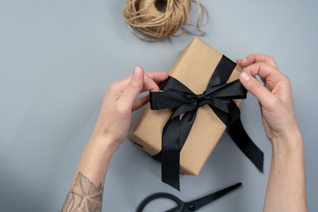 A person wraps a black ribbon around a brown box.