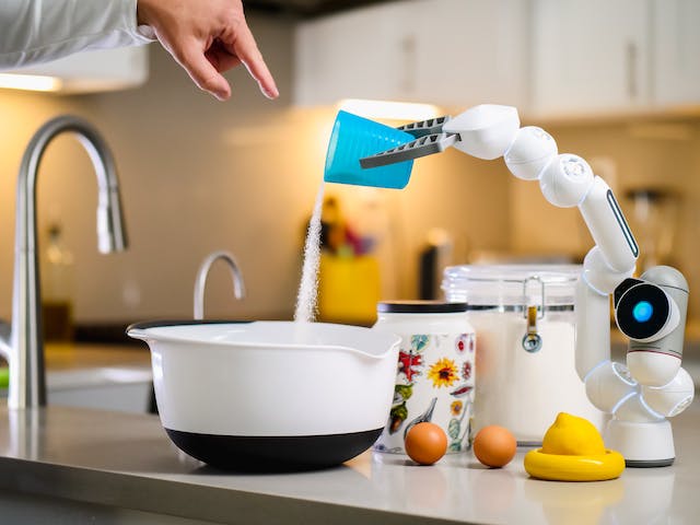 A photo of a small bot mixing ingredients while a person gives it instructions. 