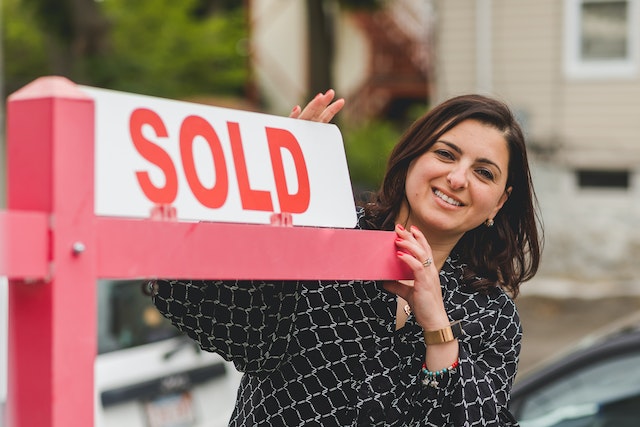 A woman stands beside a sign that says, “Sold.”
