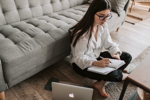 A woman sits on the floor in front of a laptop and writes a draft in a notebook.