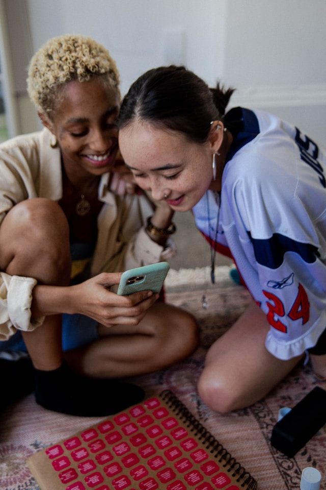 Two young women sitting on the floor, smiling and looking at TikToks on a smartphone.
