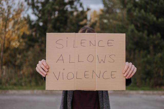 Iemand houdt een kartonnen bord omhoog met de tekst: "Zwijgen staat geweld toe.