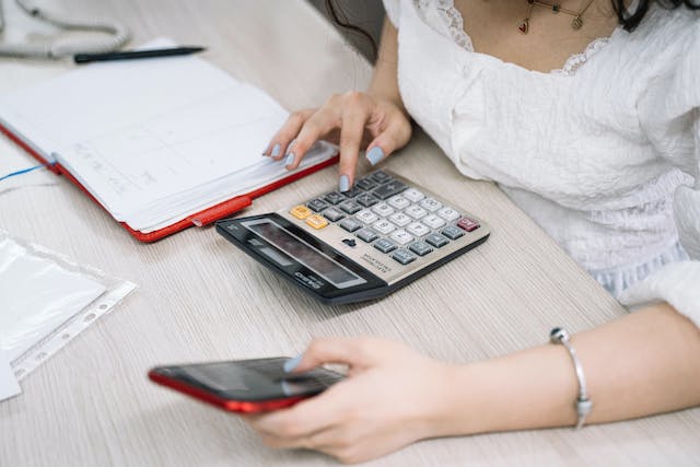 A woman checks her phone while doing calculations on a calculator. 