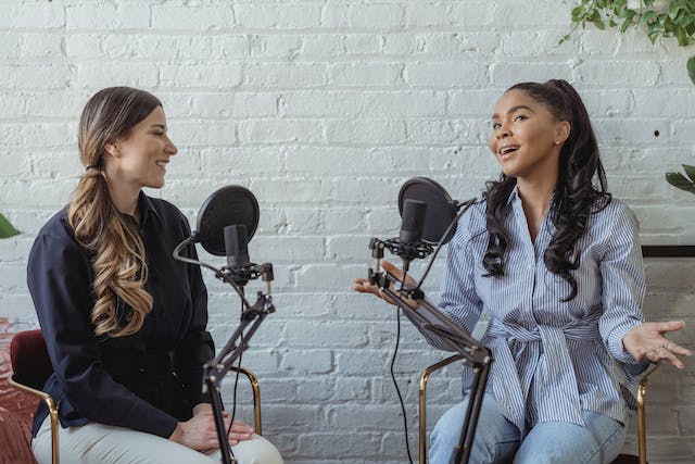 Duas mulheres sentam-se em frente a microfones para uma entrevista. 