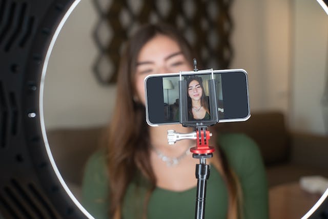 A woman sits in front of a ring light and her phone camera for a TikTok LIVE stream. 