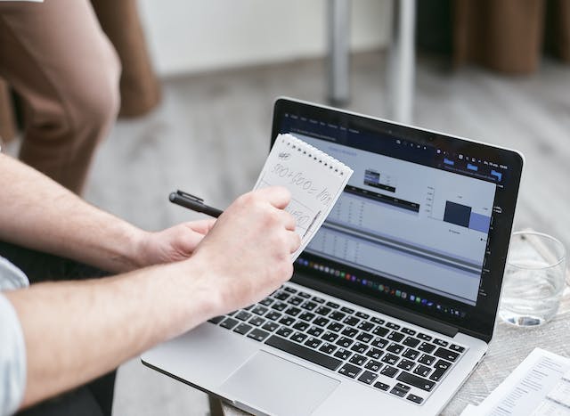 Eine Person schaut sich Diagramme auf einem Laptop an, während sie Berechnungen auf Papier durchführt. 