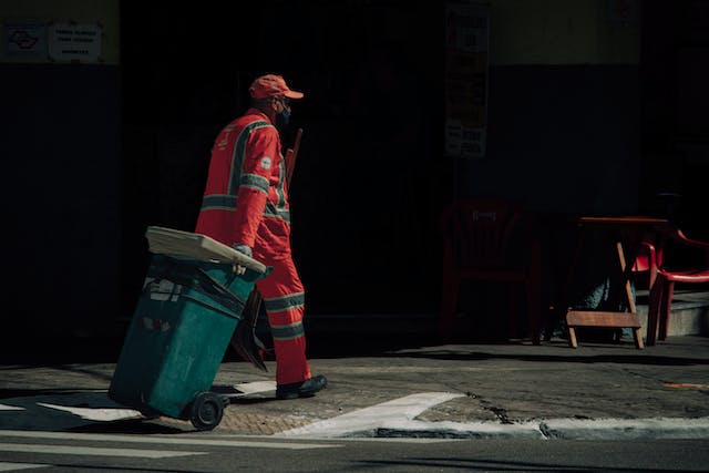 A person in red overalls pulls a rolling trash bin. 