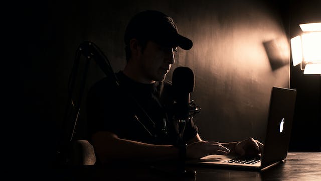 A man sits in front of a laptop and microphone for a podcast. 
