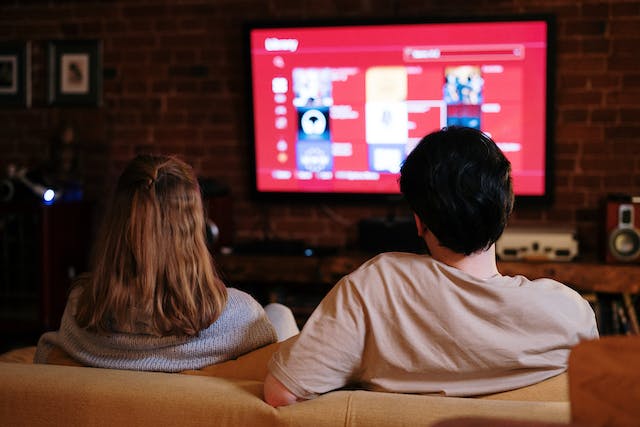 A couple sits on a couch and watches TV. 