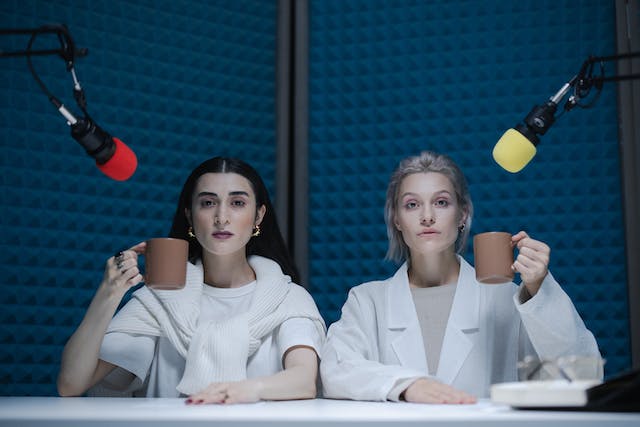 Duas mulheres sentam-se em frente a microfones em um estúdio à prova de som. 