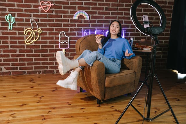 A young woman sits on a couch in front of a phone camera attached to a ring light. 