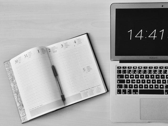  An image of an open planner with a black pen on top of the pages and a laptop displaying a digital clock. 
