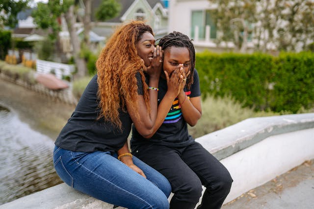 Two women sit on a bridge while whispering to each other. 
