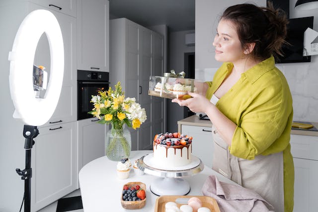 A woman in a kitchen stands in front of a ring light and a phone camera to showcase her cakes. 