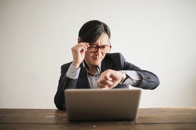 Um homem está sentado em frente ao laptop e verifica o relógio. 