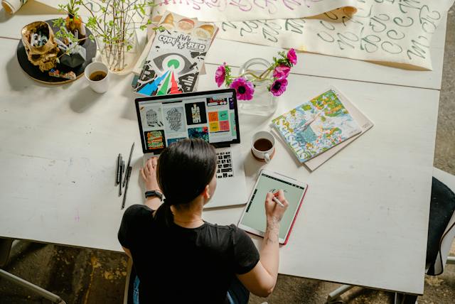Uma foto aérea de uma mulher trabalhando em seu laptop e criando arte digital em um tablet. 