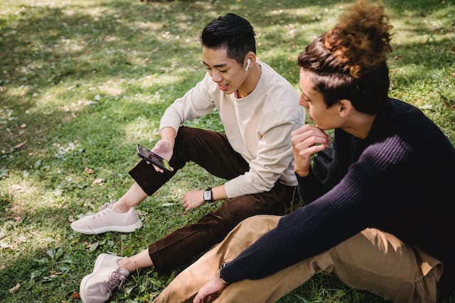 Two men sit on the grass while watching videos on a phone. 
